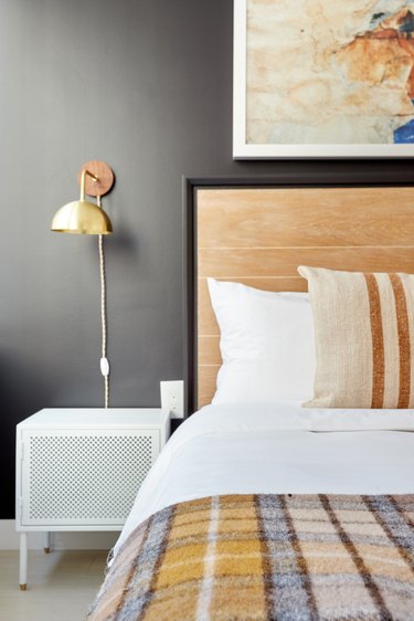 Eclectic bedroom with wood headboard, gray wall, brass sconce, white nightstand, plaid blanket.